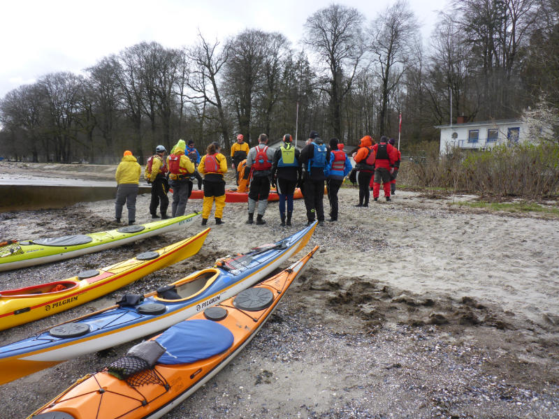 Teilnehmende stehen im Kreis am Strand, die Boote liegen an der Wasserkante